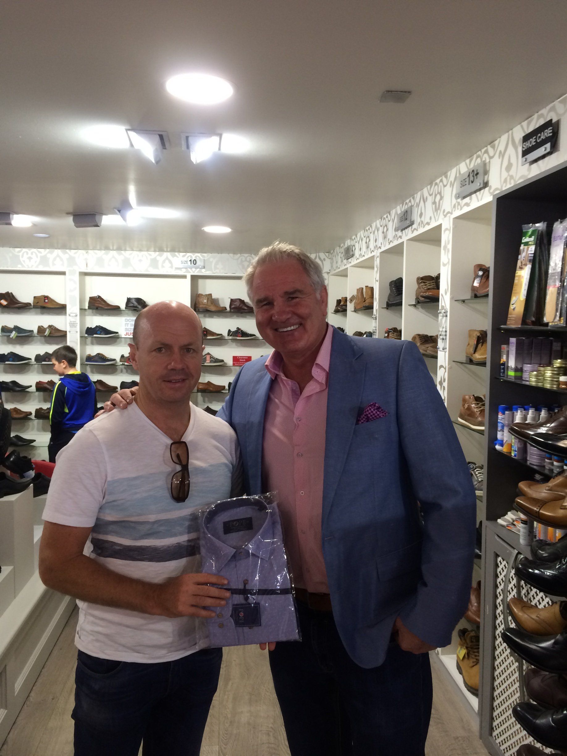 Two men pose inside a shoe store. One holds a shirt; both smile. Shelves of shoes line the walls.