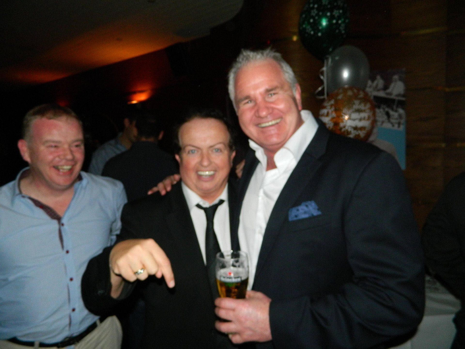 Three men smiling at a gathering; one points and holds a drink, another has his arm around the man with the drink.