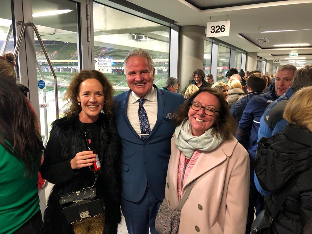 Three people smiling, posing together inside a stadium. Man in suit, two women in coats, crowd visible.