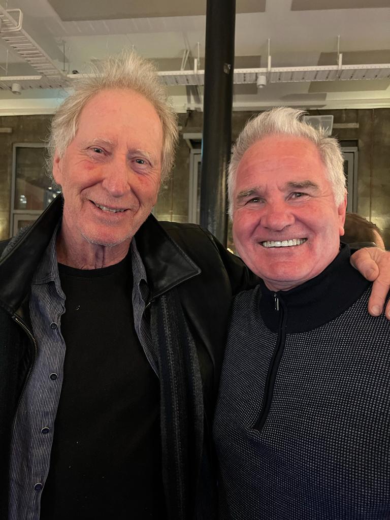 Two smiling men pose close together indoors. One man has gray hair, leather jacket. The other man is older, with gray hair and a patterned sweater.