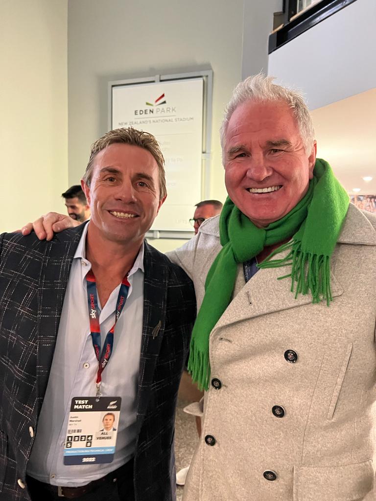 Two men smiling for the camera. One wears a blazer, the other a green scarf and coat. They are indoors.