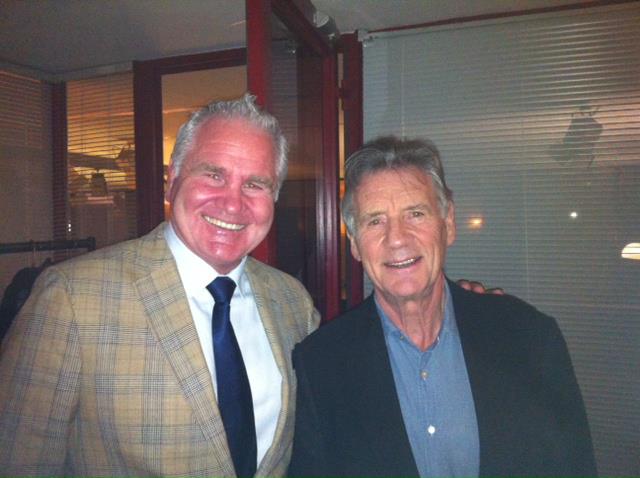 Two men smiling, one in a plaid blazer and tie, the other in a black jacket, posing indoors.