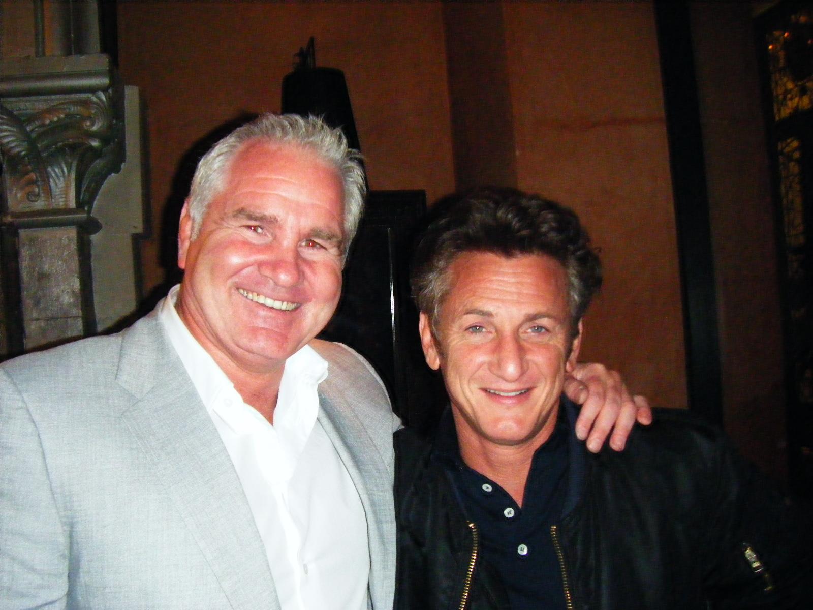 Two men smiling, posing close together. One with gray hair, light jacket, other with dark hair, black jacket. Indoor setting.