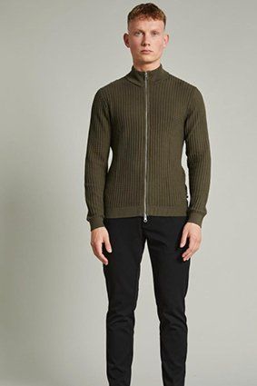 Man wearing a green zip-up sweater and black pants. Standing in a studio setting.