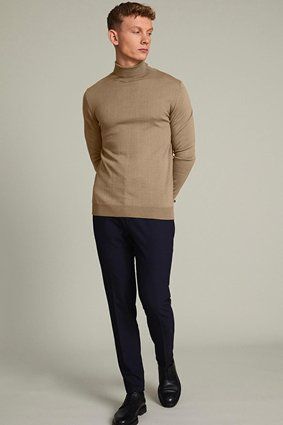 Man wearing a tan turtleneck and navy pants, standing with hands behind his back.