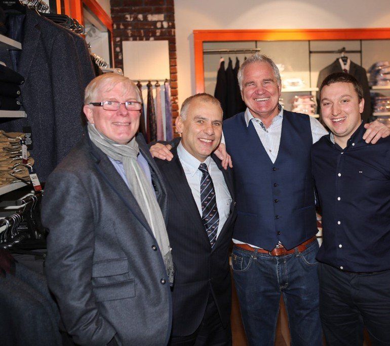 Four men smiling and posing in a clothing store, arms around each other.