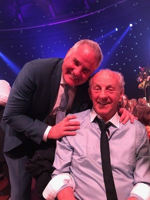 Man in suit smiles at man seated, arm around his shoulder, celebratory setting.