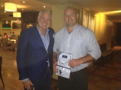 Two men posing in a restaurant; one holding a shirt. The man on the left wears a blue blazer. The other wears a light blue shirt.