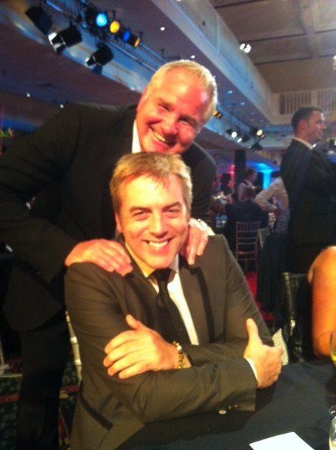 Two men in suits smiling, one behind the other. The man behind has his hands on the other man's shoulders. Party setting.