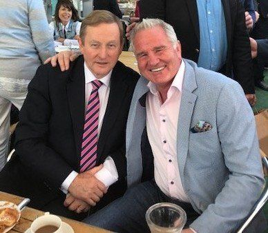 Two men smiling, one in a suit with a pink striped tie, the other in a blue blazer. Outdoor setting.