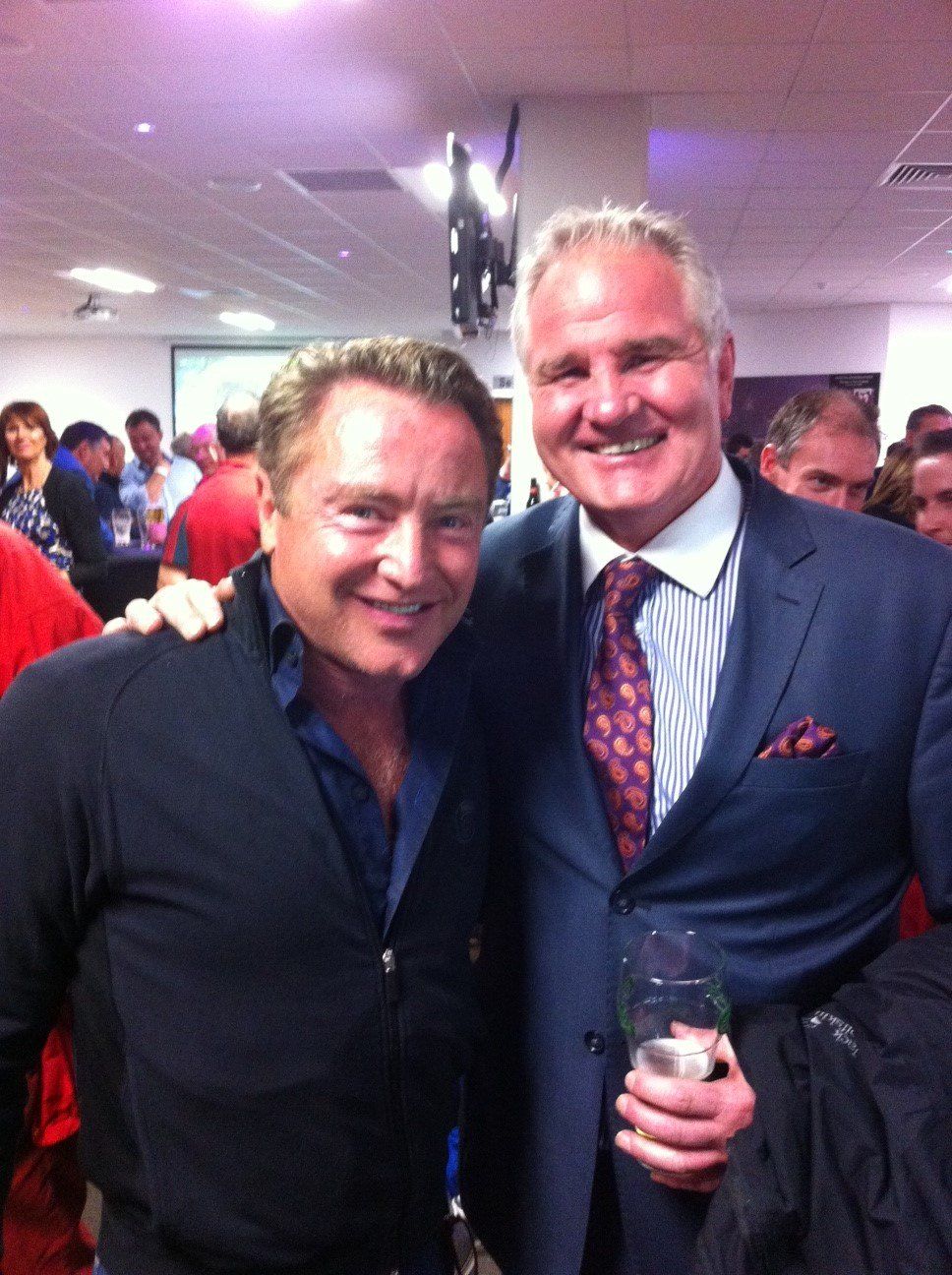 Two men smiling, arm around each other. One in suit, holding a drink; other in black zip-up.