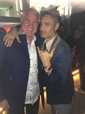 Two men smile, pose close together. Man on left has arm around the other, both in suits, one shows a hand gesture.