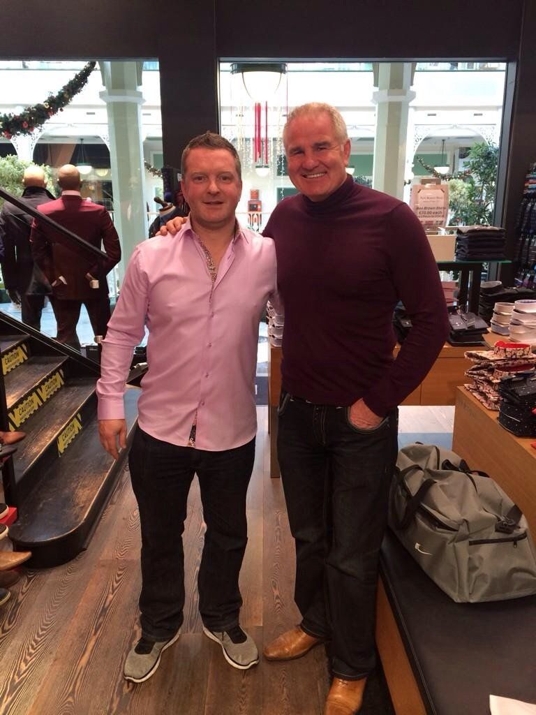 Two men pose for a photo inside a clothing store. One wears a pink shirt, the other a burgundy sweater.