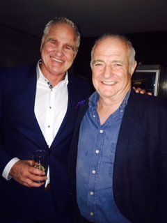 Two men smiling, one in a blue suit, the other in a blue shirt, both in a dark setting.