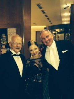 Three people in formal attire pose for a photo. A woman in a black dress stands between two men in tuxedos, smiling.