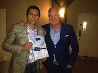 Two men smiling, one holding a packaged white dress shirt, both wearing blazers, indoors.