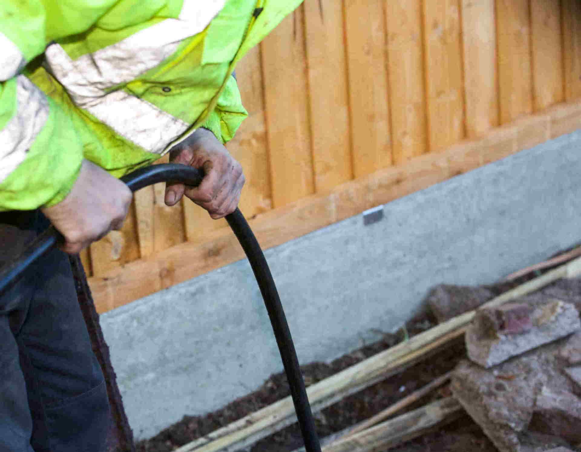 Worker in a lime safety jacket holding a black hose beside a wooden fence and concrete foundation.
