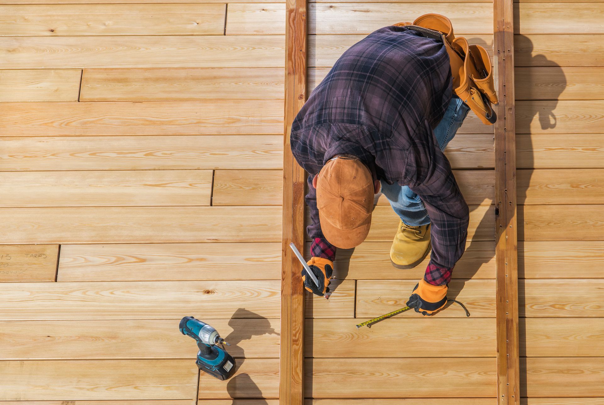 Person measuring wood planks with tools, wearing a hat, leaning over, working on construction.