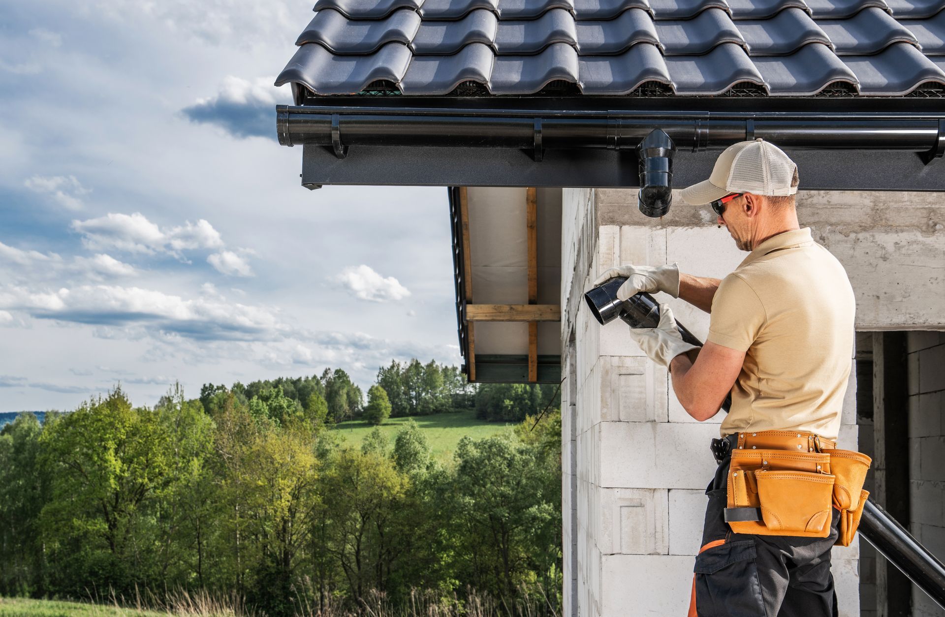 Man installing a gutter on a white building; green trees and blue sky in the background.