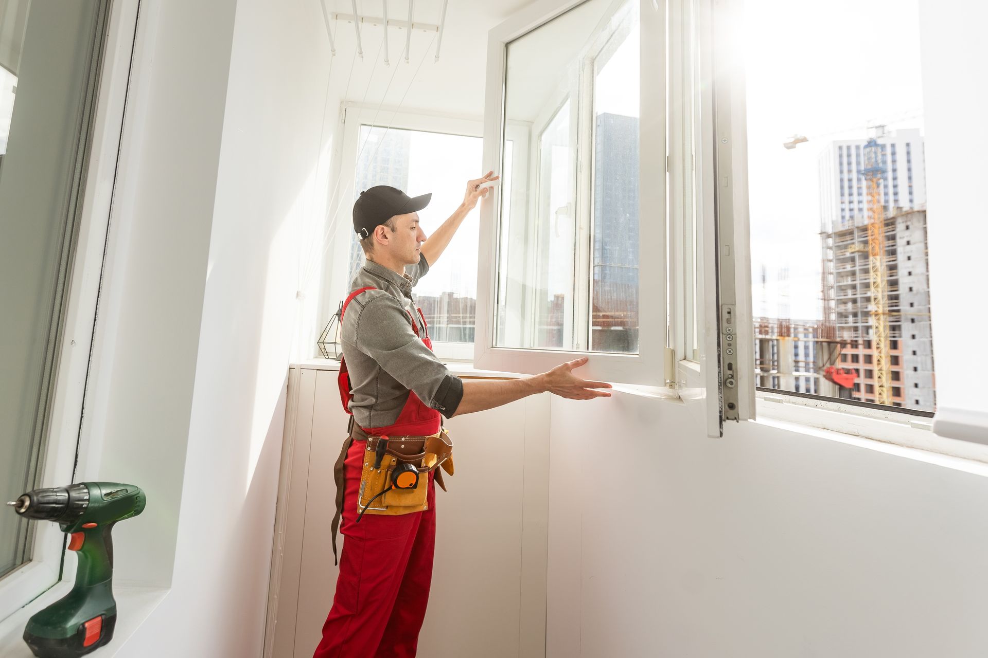 A handyman in red overalls installs a white window in a bright room with a city view.