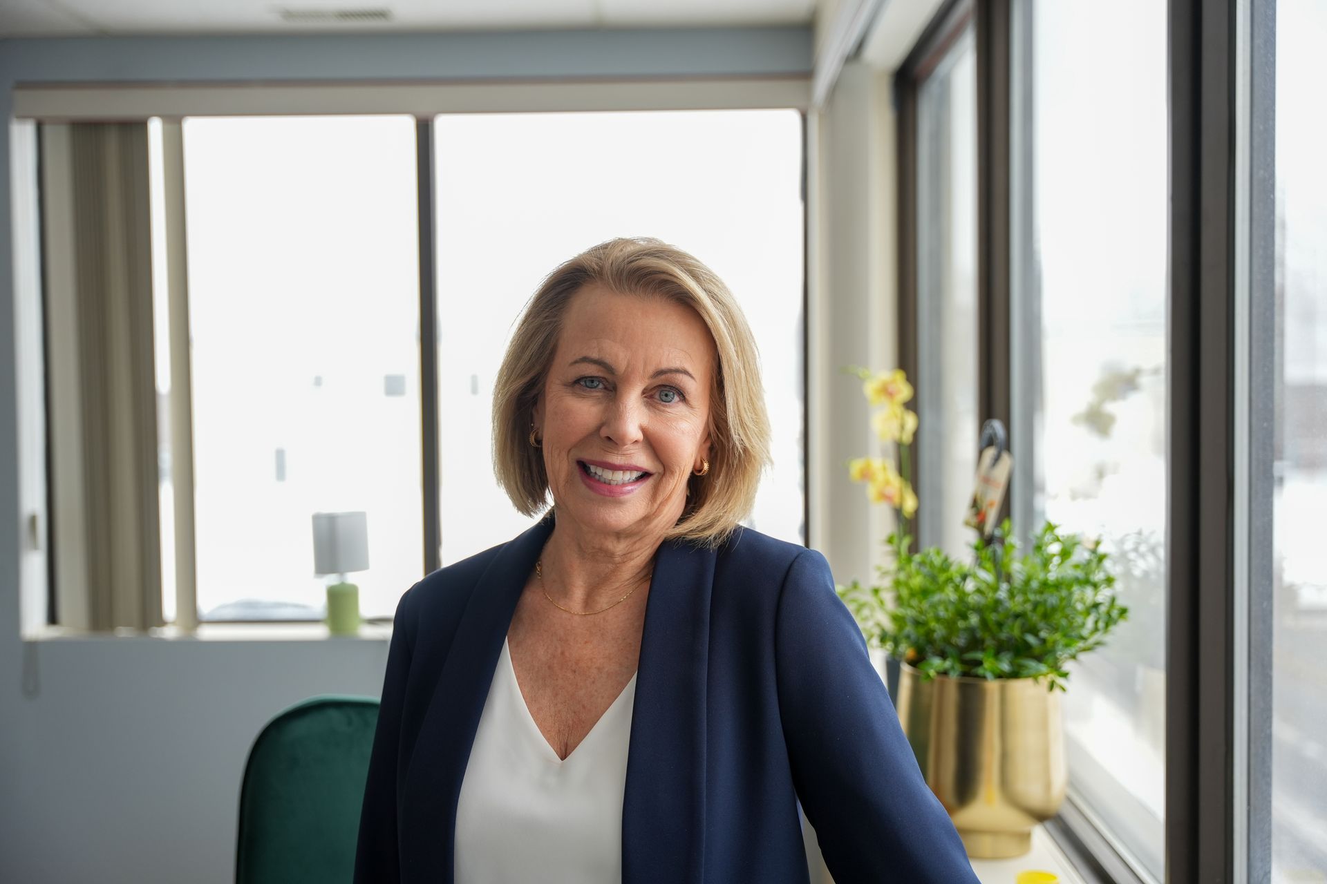 A smiling older woman in a blue blazer and white top stands near a window, indoors.
