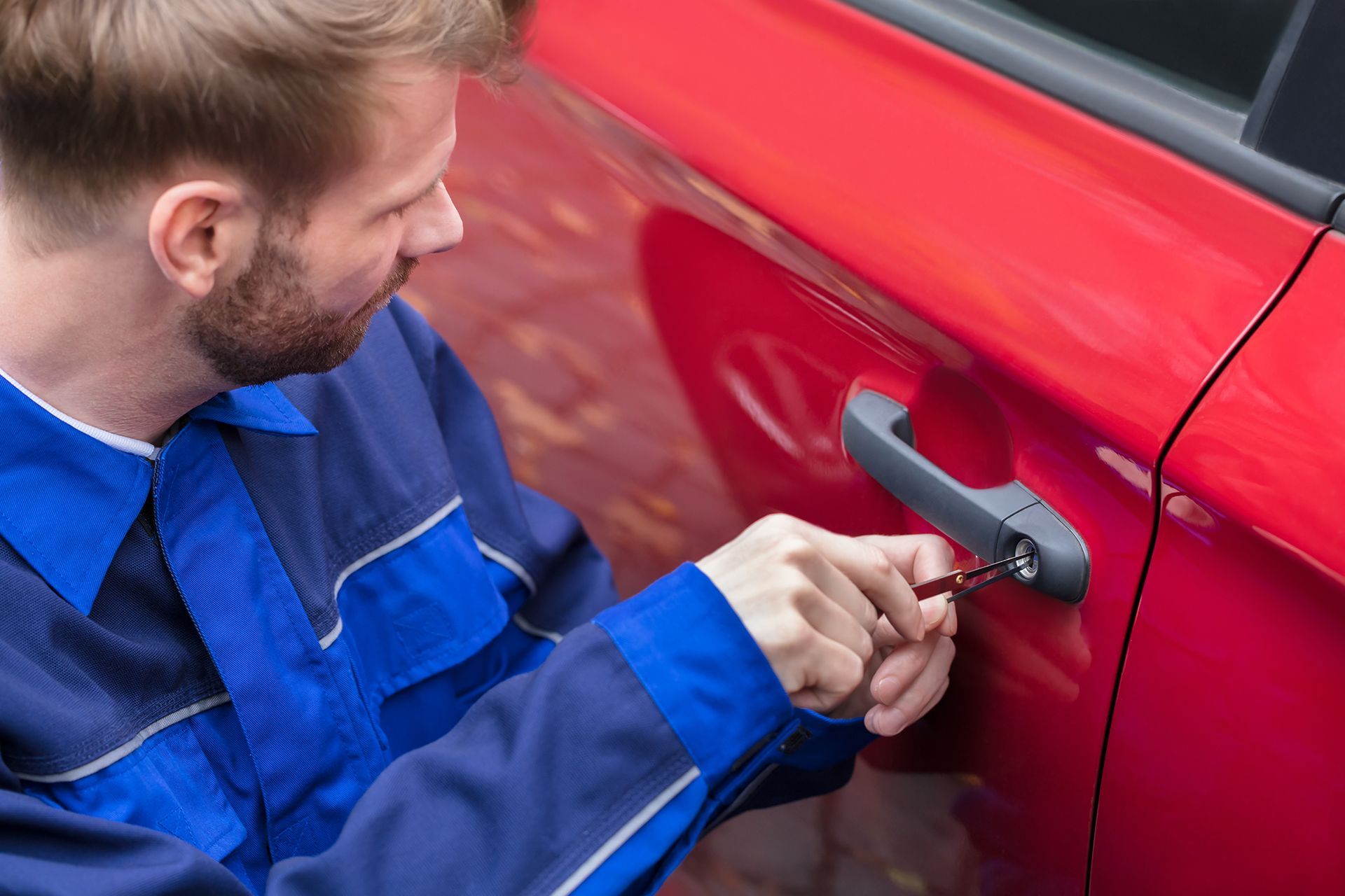 A man is using a screwdriver to open the door of a red car.