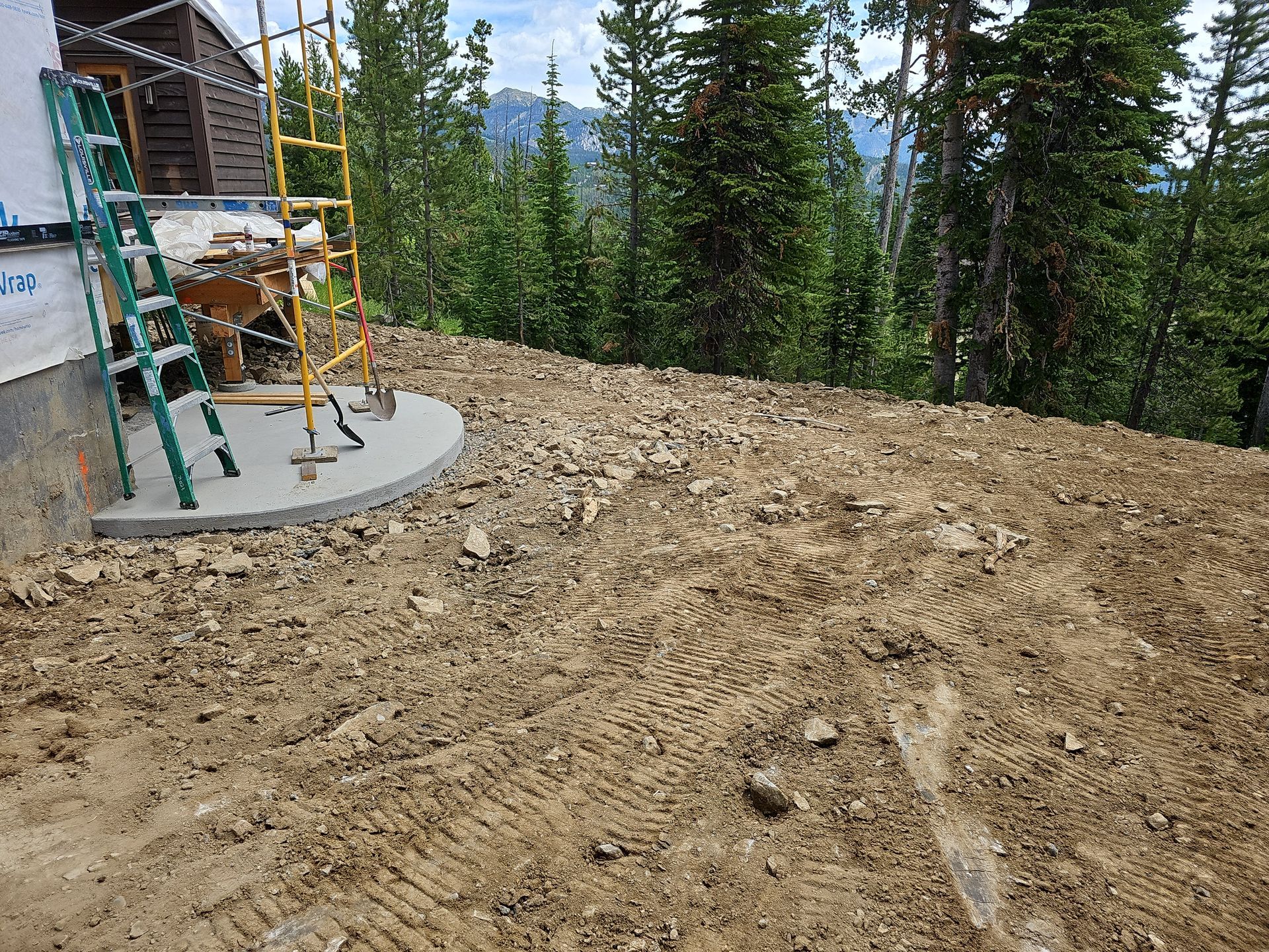 Construction site: dirt ground, concrete base, scaffolding, ladder, trees, mountains.
