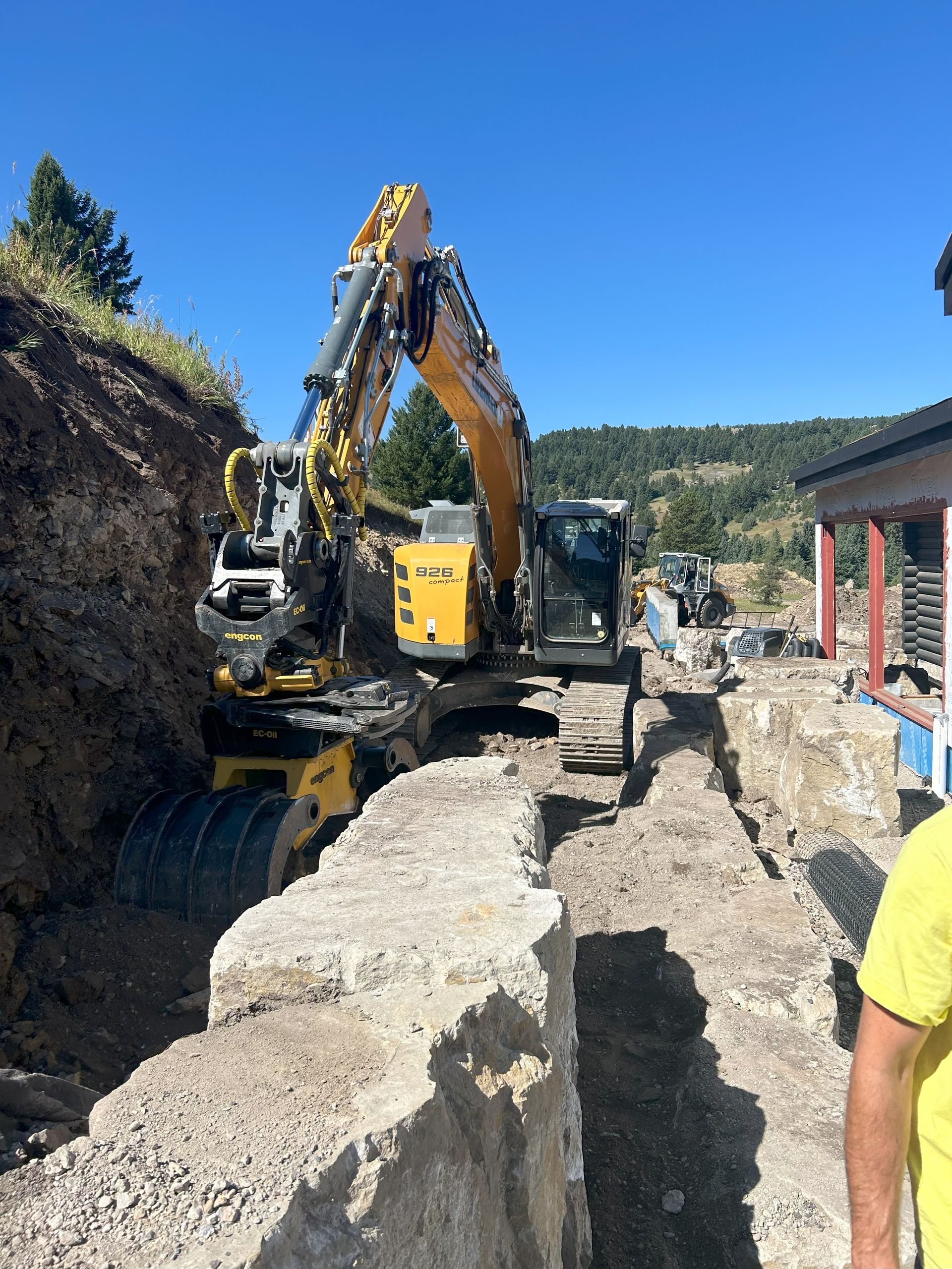 A man in a yellow shirt stands in front of an excavator