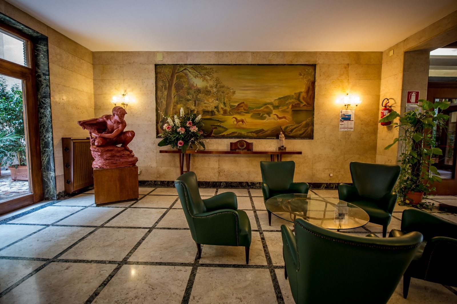 Un atrio con sedie verdi, un tavolo, una statua e un grande dipinto sul muro.