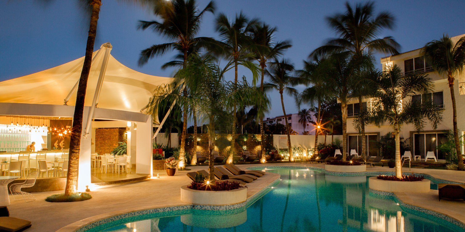 Piscina de H2Otel by LD iluminada al anochecer, rodeada de palmeras y áreas de descanso, invitando a la relajación.