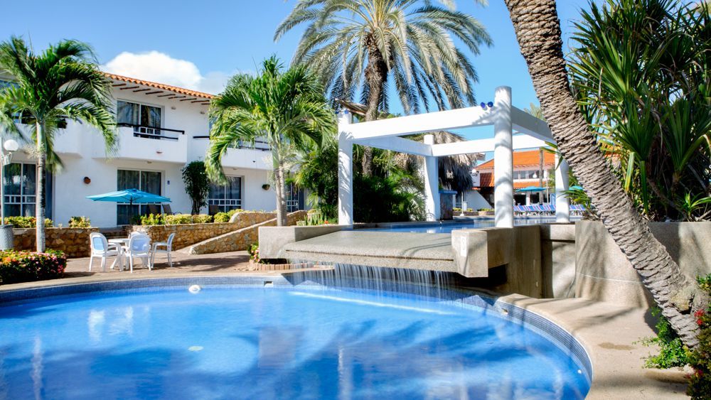 Piscina curva con cascada en LD Palm Beach, Playa El Agua, rodeada de palmeras y gazebo blanco.