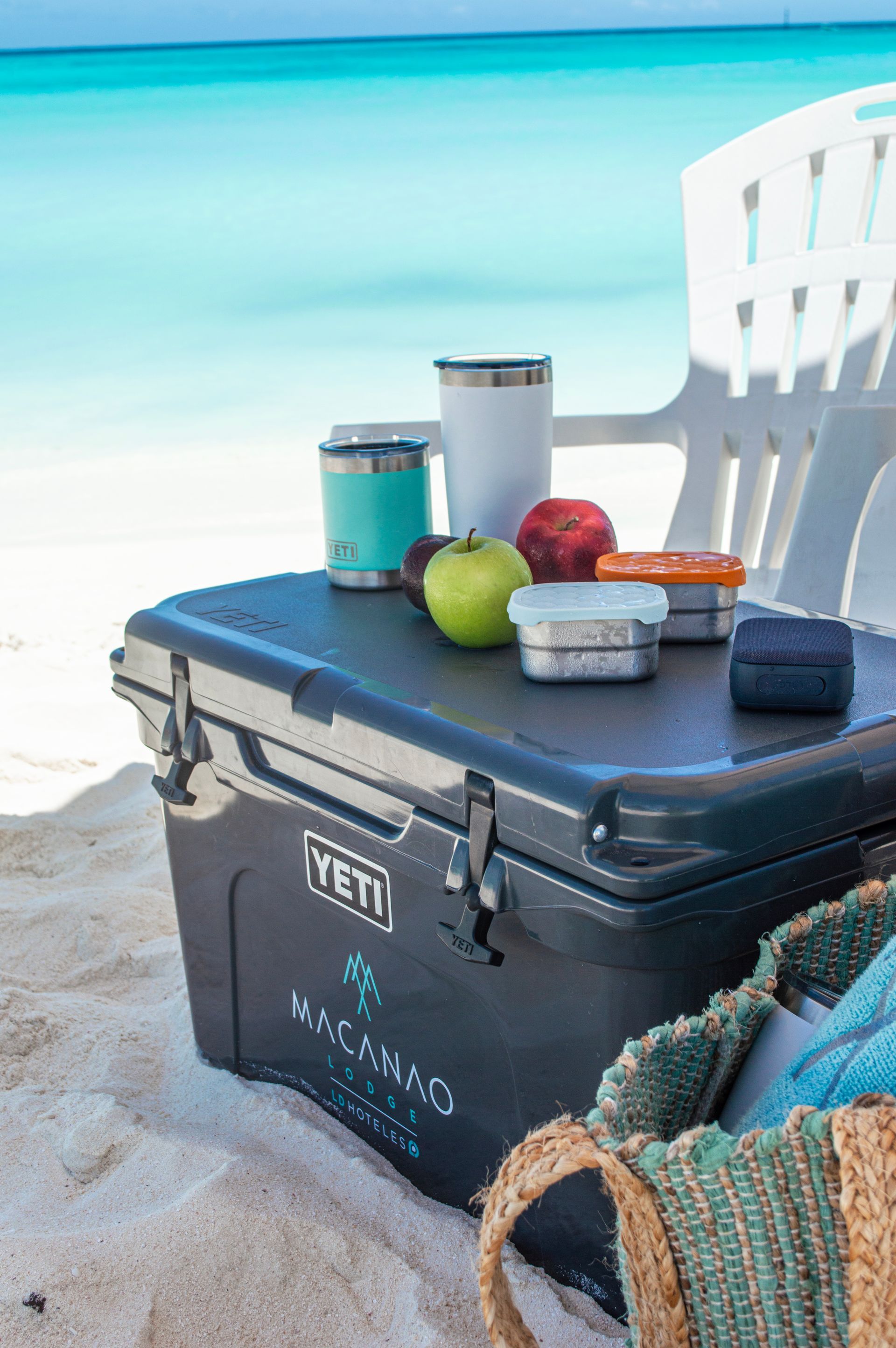 Cava YETI oscura con tumblers y manzanas sobre arena en playa, ideal para día soleado en Los Roques.