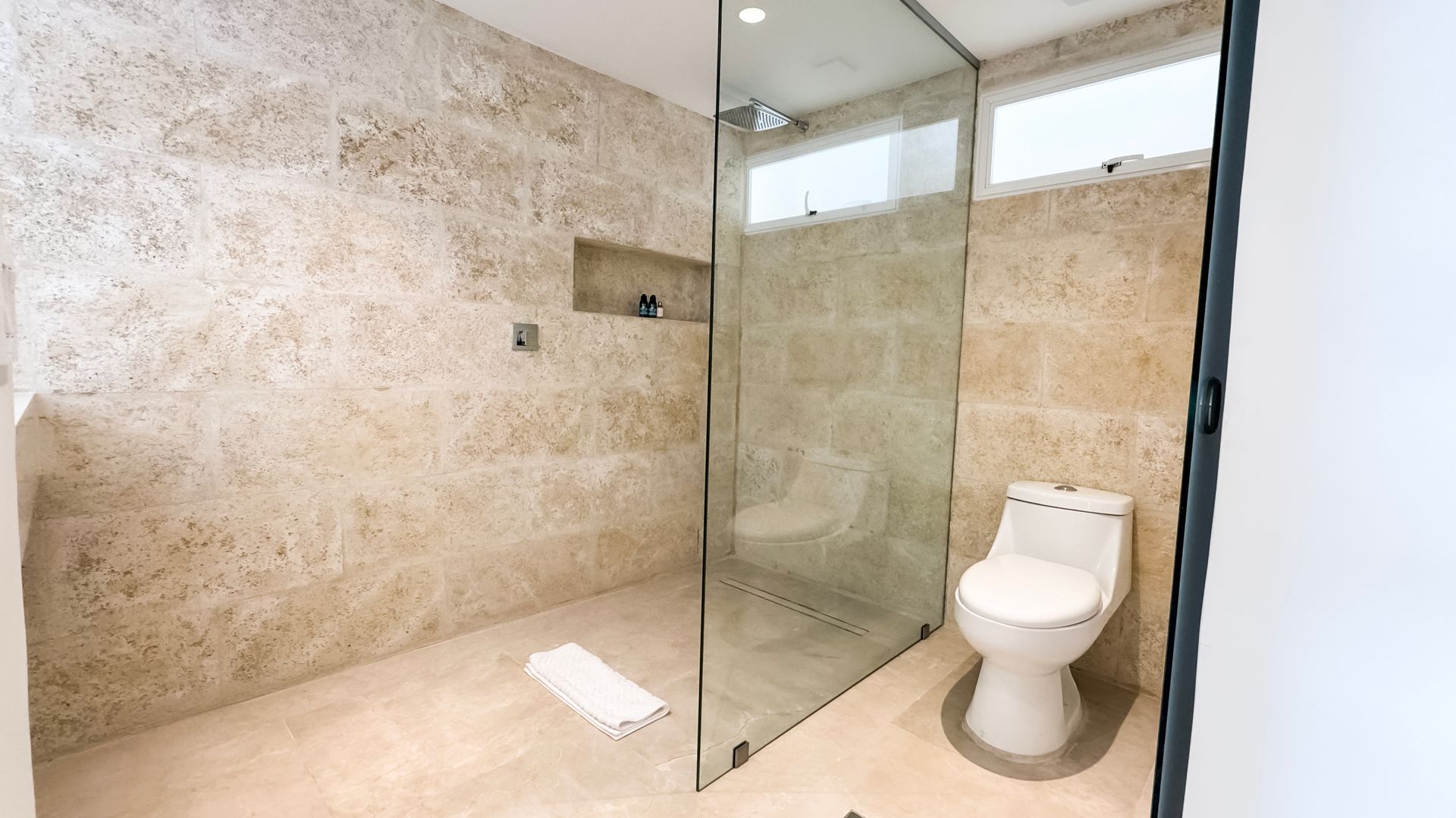 Baño de hotel en Los Roques con azulejos beige, ducha de cristal y toallas blancas, ofreciendo un ambiente limpio y moderno.