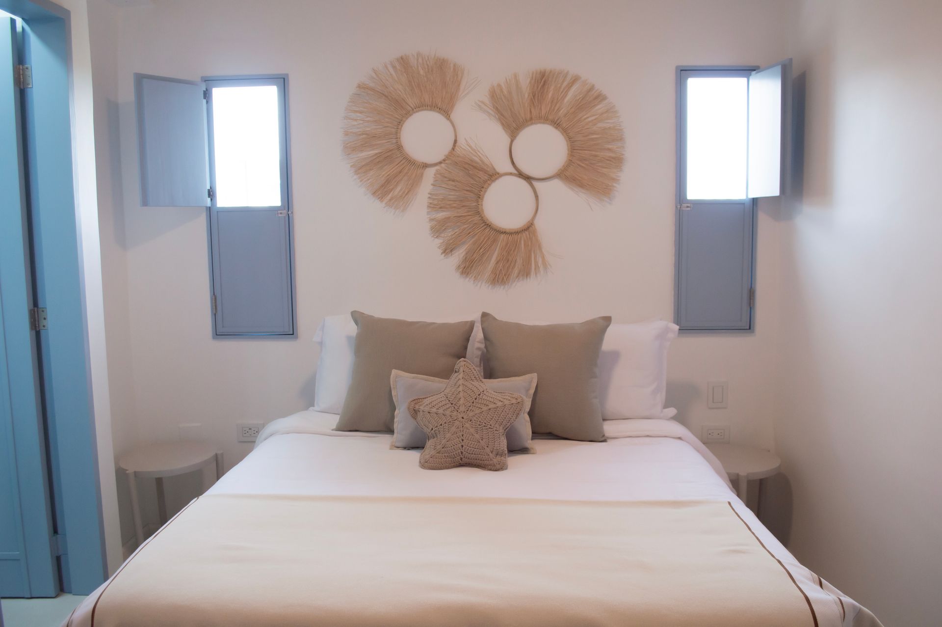 Habitación luminosa con cama doble, decoración en tonos blancos y beige, y ventana con contraventanas azules.