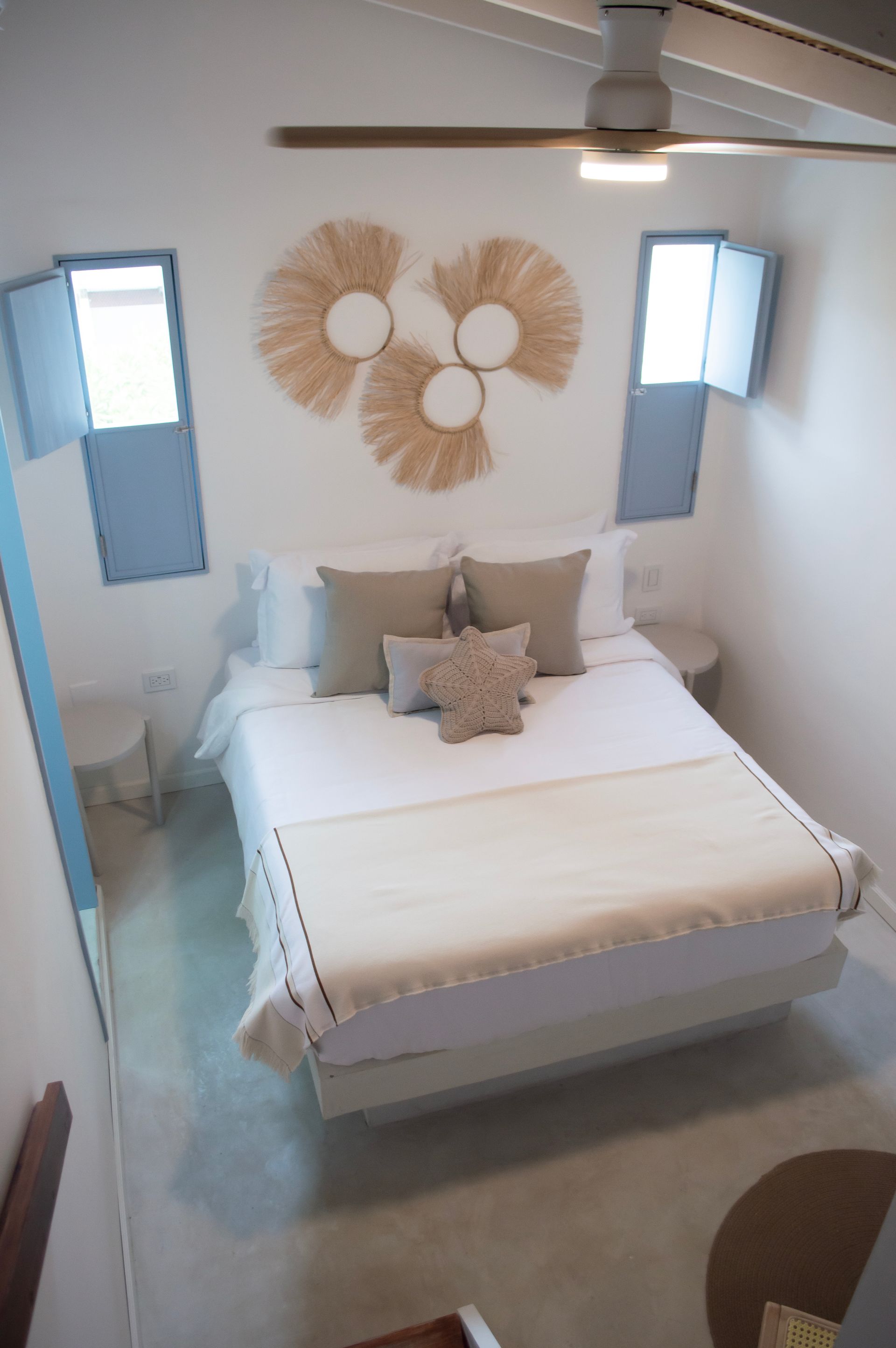 Habitación luminosa con cama doble, decoración en tonos blancos y beige, y ventana con contraventanas azules.