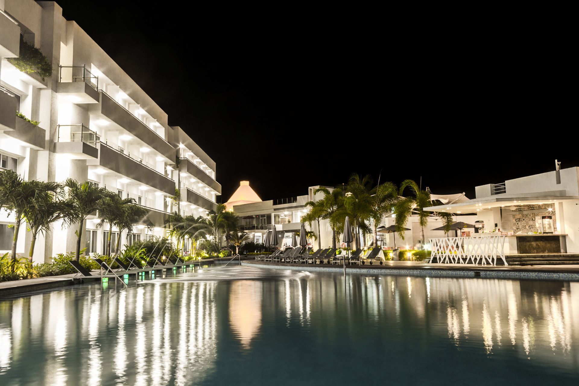 Piscina iluminada de LD Plus, Playa El Agua, rodeada de palmeras y edificio del hotel.