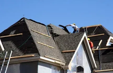 Workers repairing a dark shingled roof on a house with a white dormer.