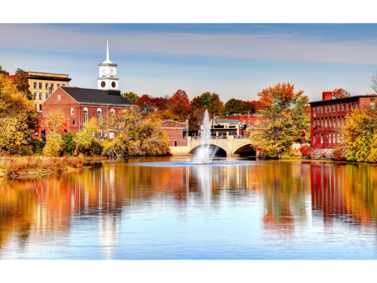 A picturesque autumn scene: brick buildings and vibrant fall foliage frame a reflective lake with a central fountain, under a blue sky.