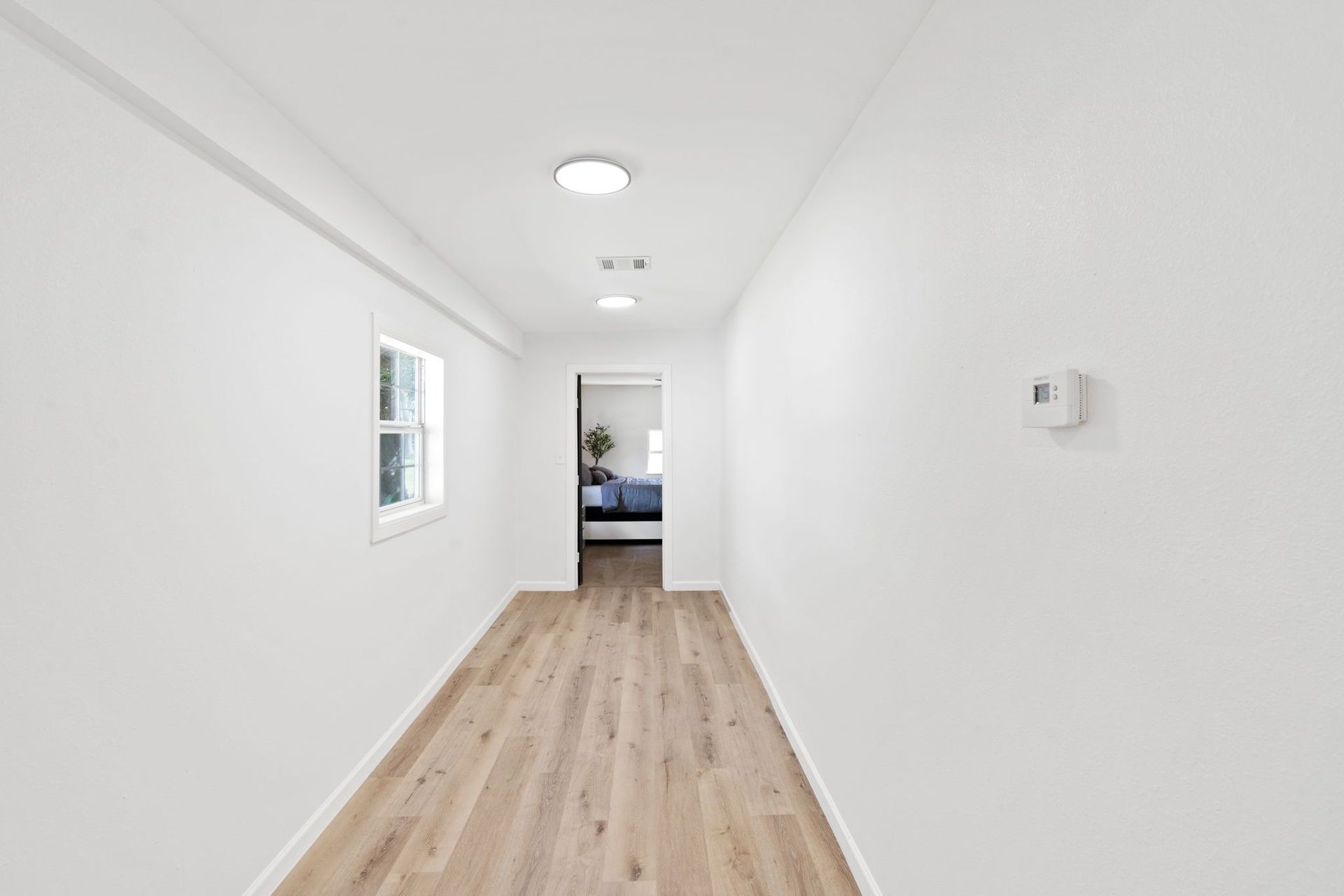 An empty hallway with hardwood floors and white walls leading to a bedroom.