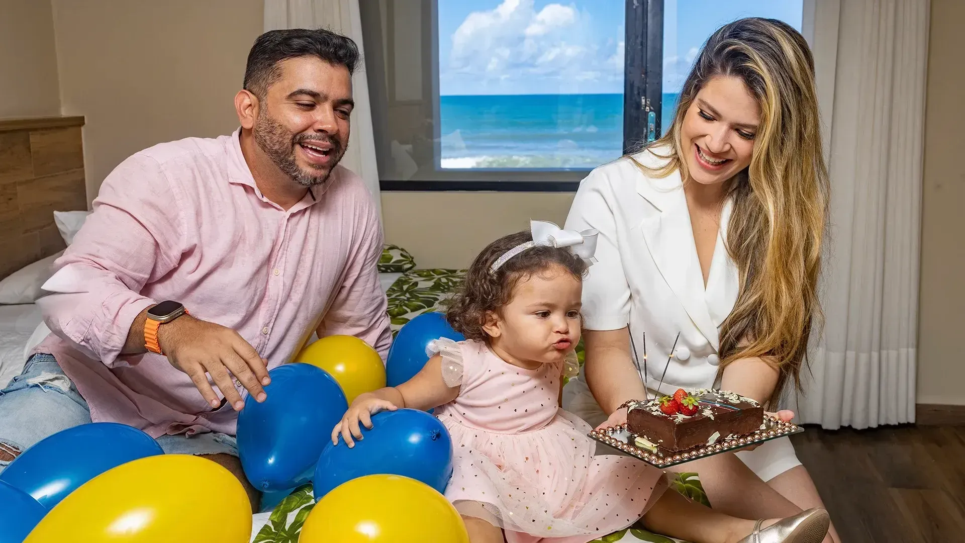 Um homem e uma mulher estão sentados em uma cama com uma menina segurando um bolo.