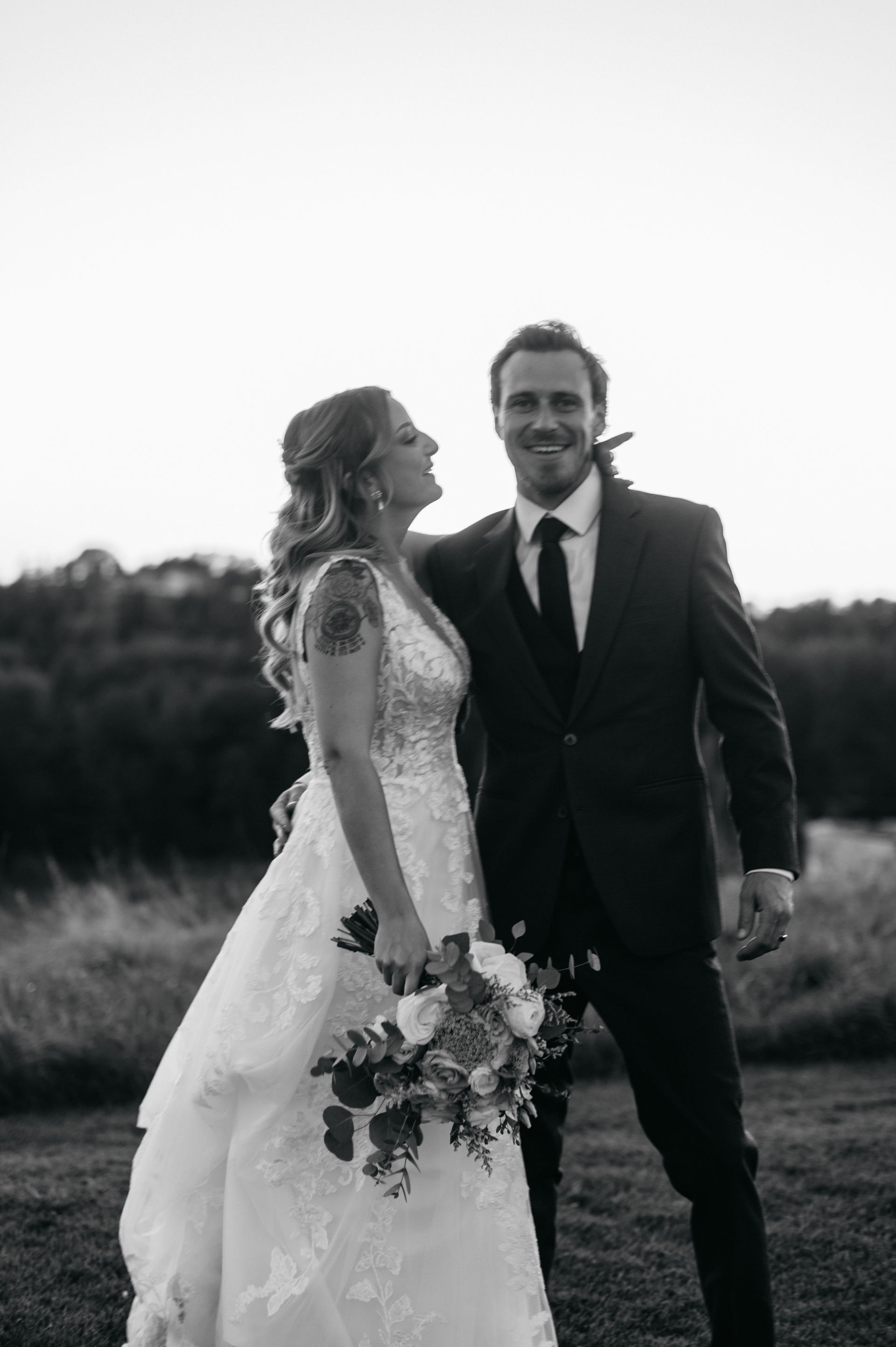 Bride and groom smiling, embracing, outdoors. Bride in lace dress, holding flowers; groom in suit.