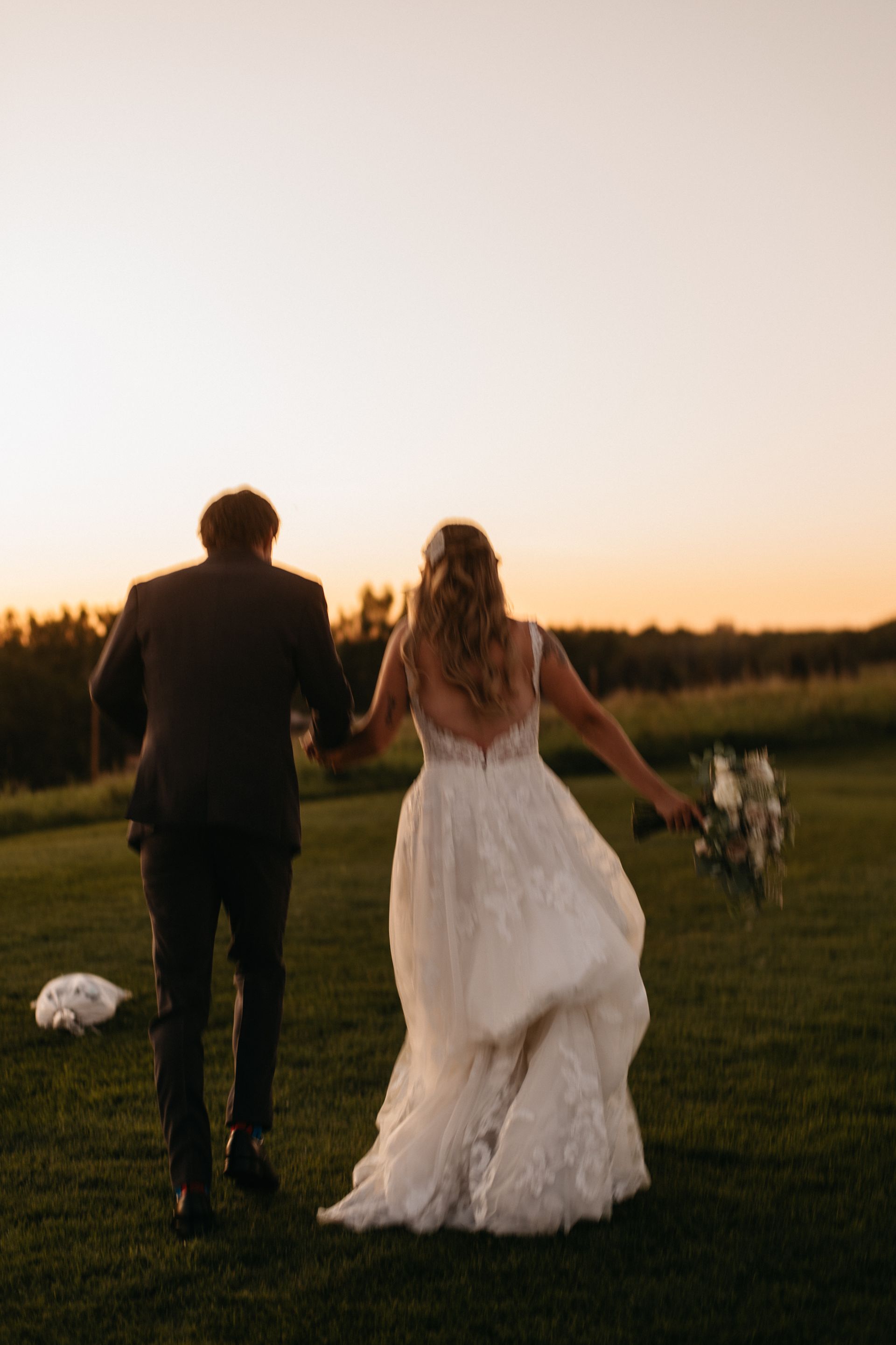 Newlyweds run, holding hands, across a grassy field at sunset. Bride in white dress carries a bouquet.