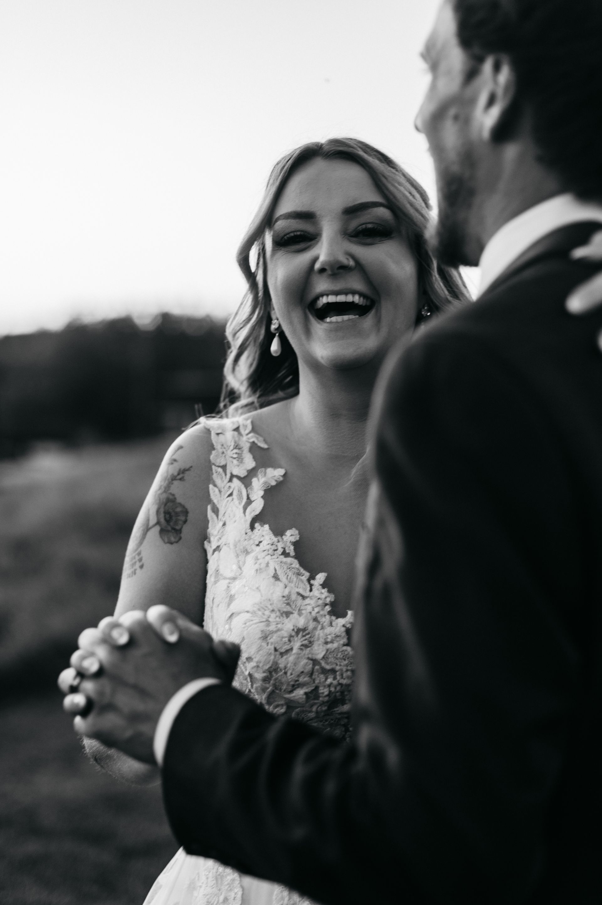 Bride laughing, dancing with groom. Outdoor setting, wearing a lacy wedding dress, holding hands, black and white photo.