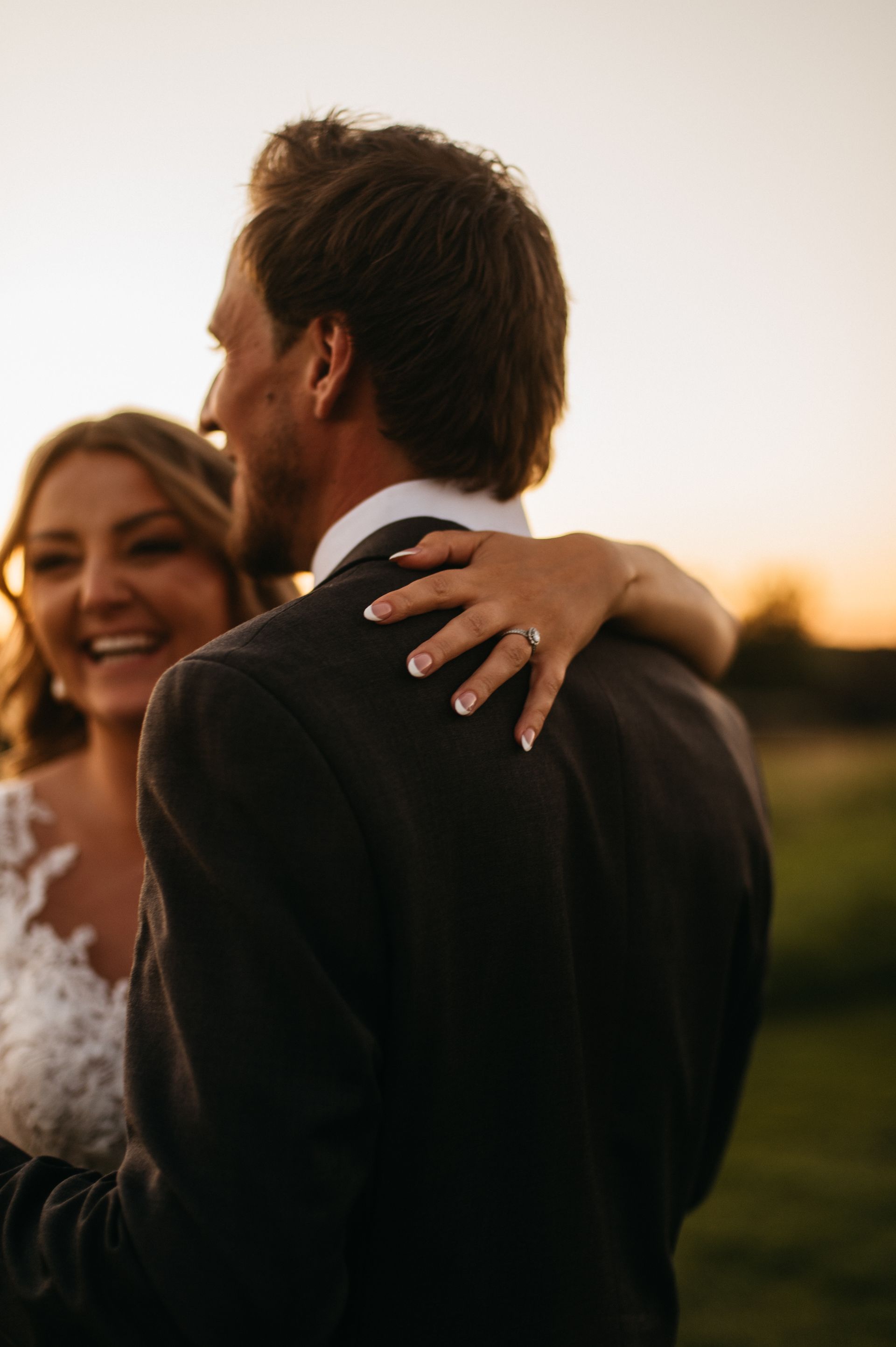 Bride smiles, hugging groom from behind. Sunset in the background. She wears a lace dress. Ring is visible.