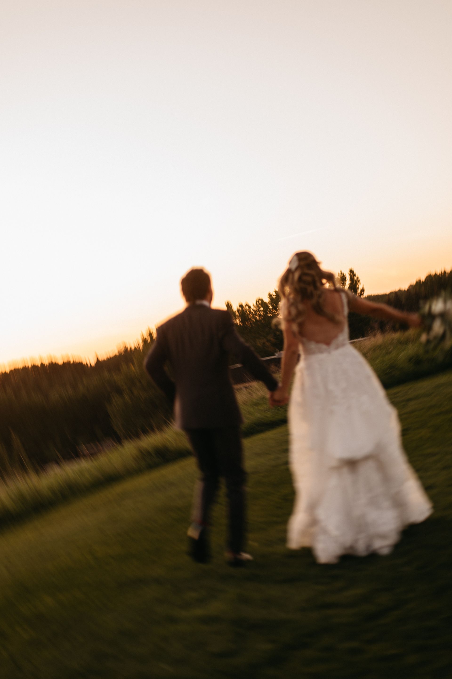 Couple running hand-in-hand across a grassy field at sunset. The bride wears a white dress.
