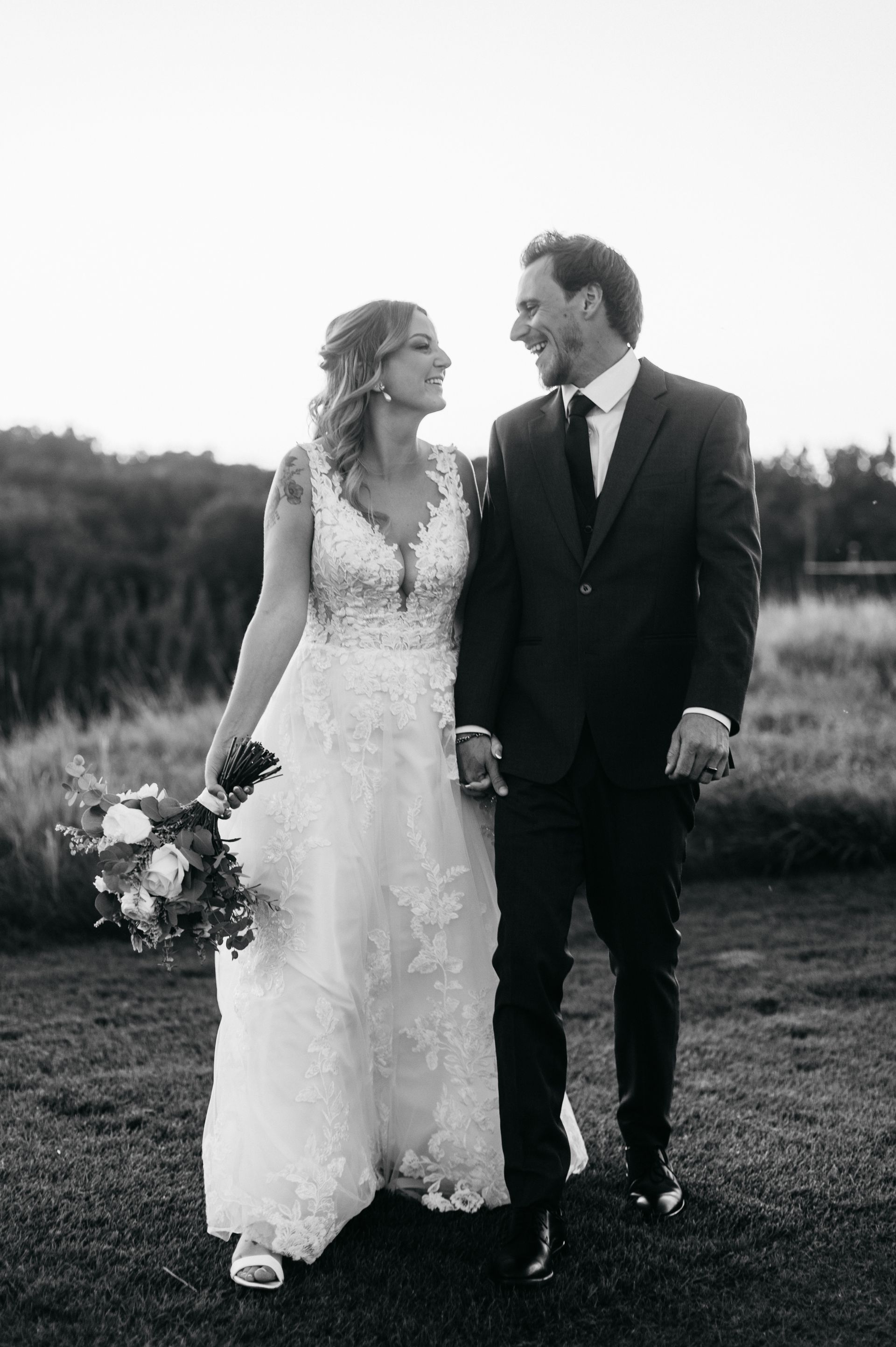 Bride and groom holding hands, smiling at each other, walking in grassy field.