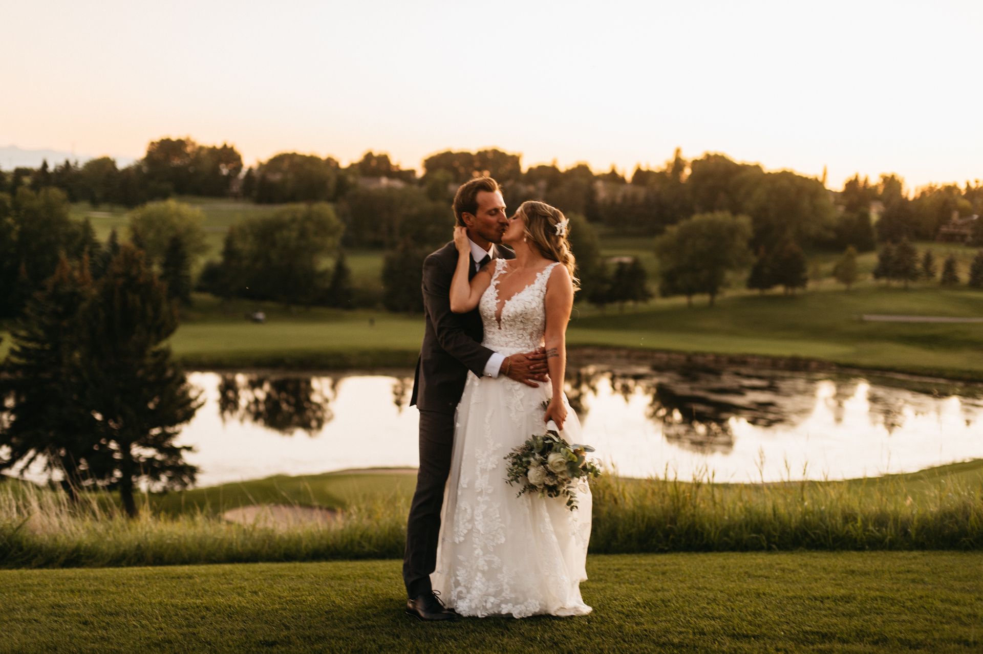 Newlyweds embrace on a golf course at sunset, lake in background.