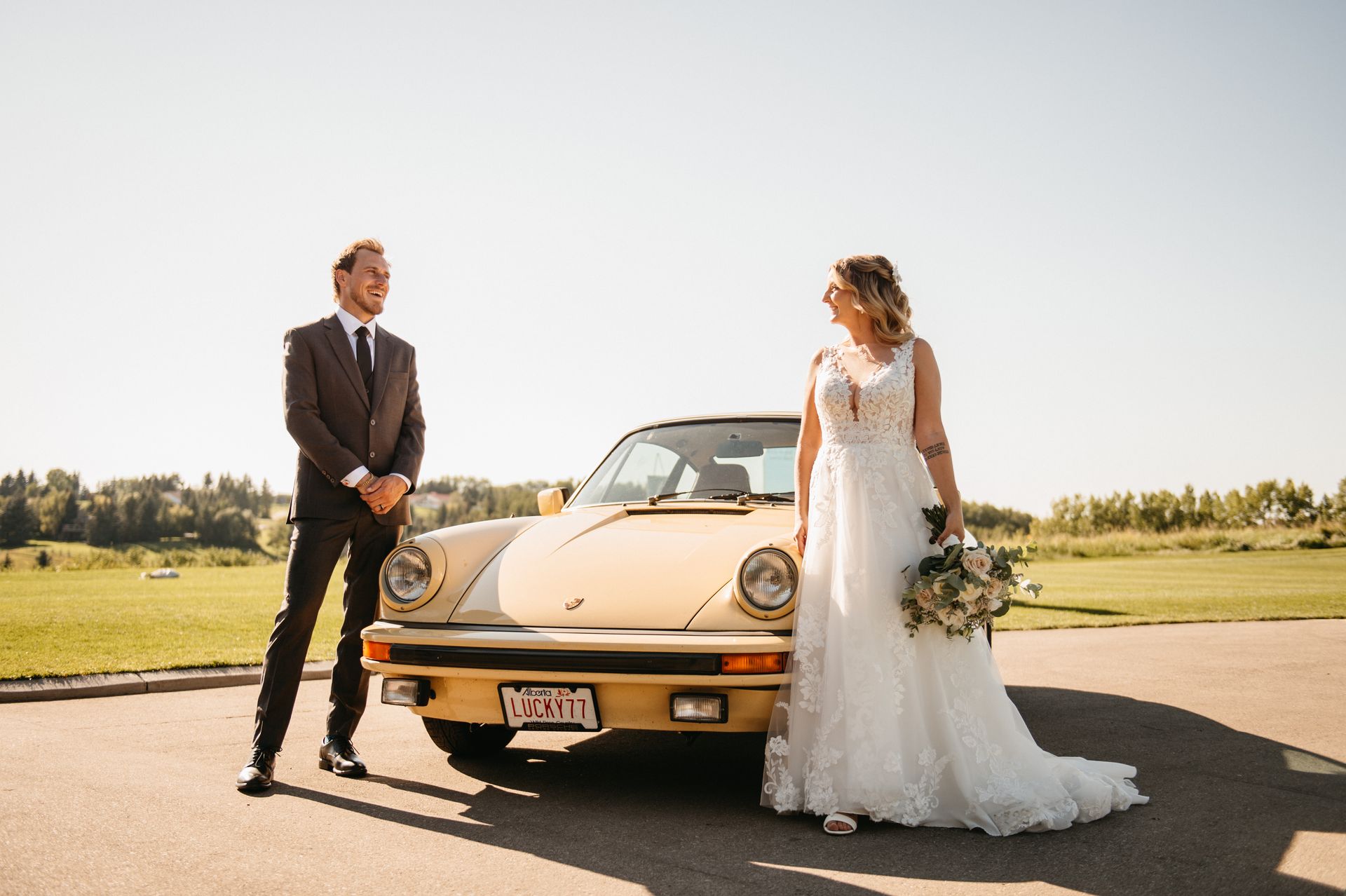 Newlyweds pose beside a vintage yellow Porsche on a sunny day. The groom laughs, the bride smiles.
