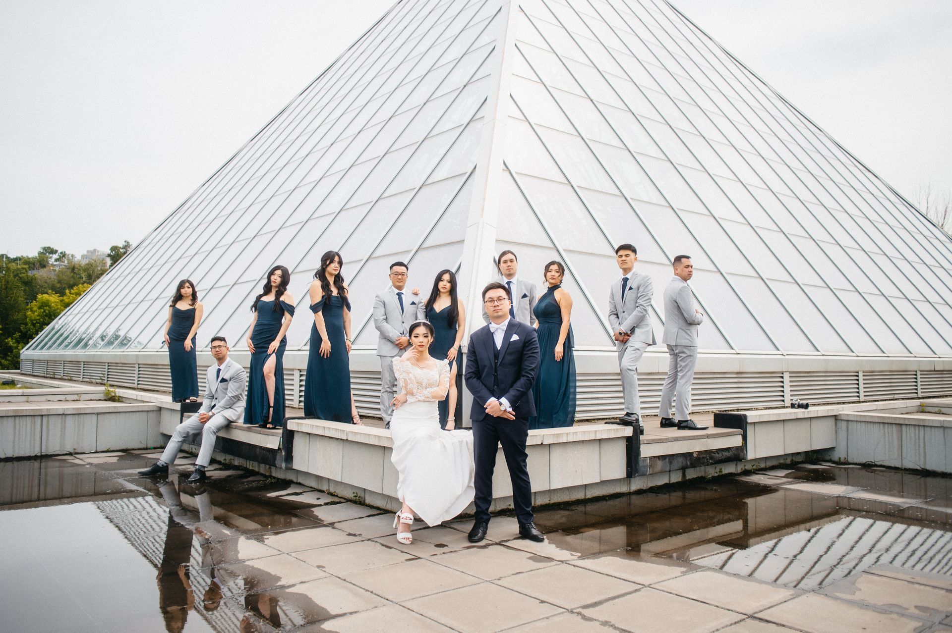 Wedding party poses in front of a glass pyramid. Bride and groom in focus, others in formal wear. Cloudy sky.
