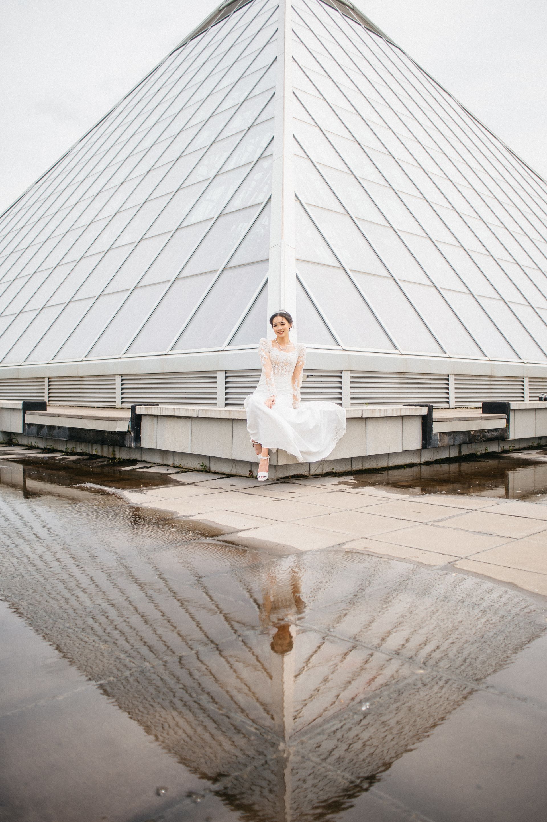 Woman in white dress sits on ledge, pyramid building behind her, reflection in water.